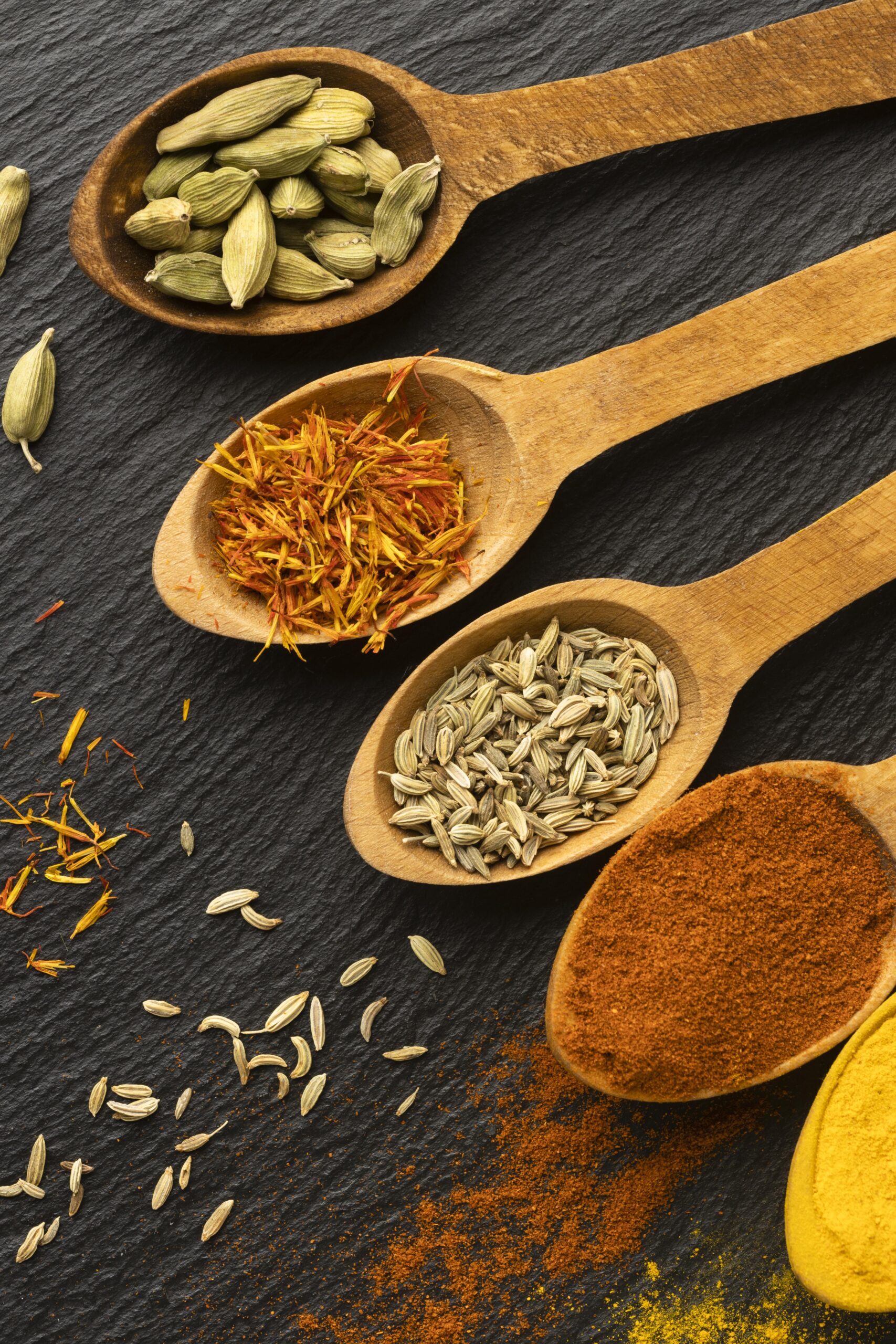 Premium Indian spices including cardamom, fennel seeds, safflower petals, and chilli powder displayed in wooden spoons on a dark background.